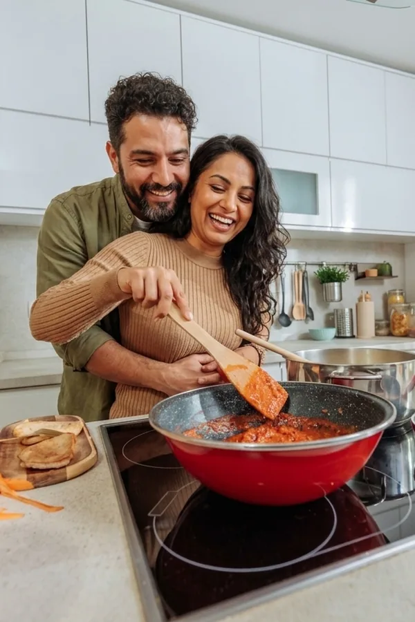A couple laughing while stirring a pan of sauce on the hob.